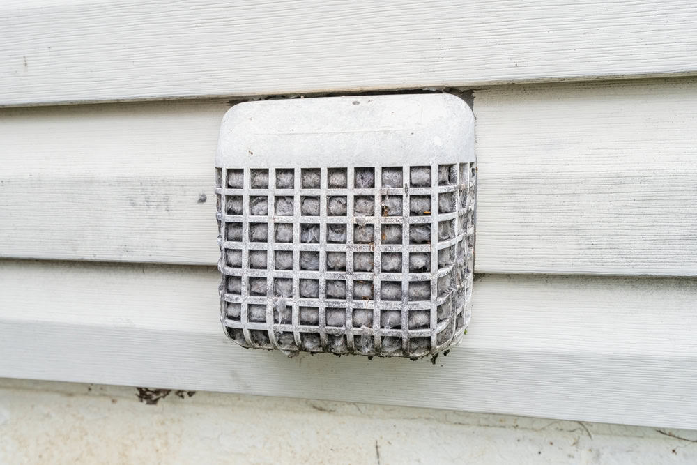 The vent cover on the house, featuring a white rectangular grid design and attached to light-colored exterior siding, shows significant lint buildup. For optimal home safety and efficiency, consider scheduling a dryer vent cleaning in Sacramento.
